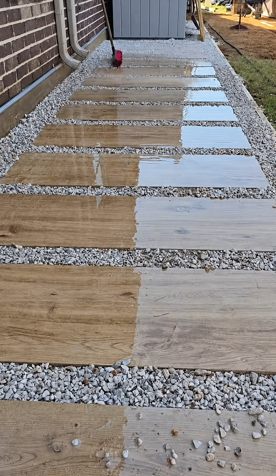 A side-yard walkway with alternating light and dark wood-look stepping-stone pavers set in white and gray pea gravel, with wet areas reflecting light, bordered by brick house wall on the left and blue metal shed on the right.
