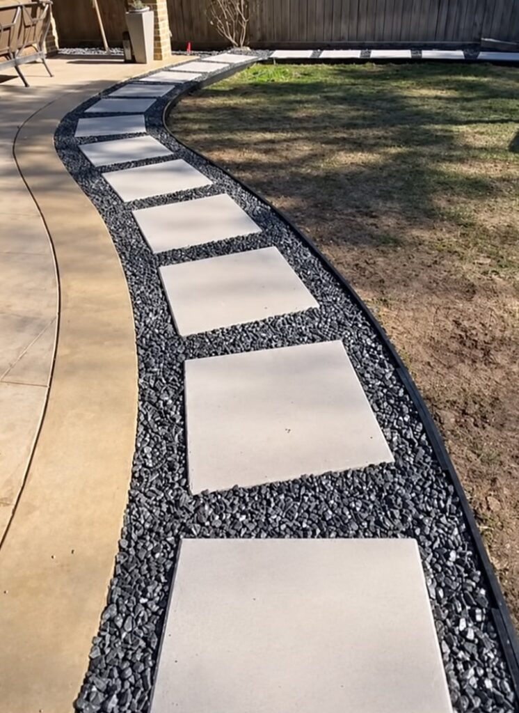 Curved concrete square stepping-stone walkway with dark gray gravel borders and black landscape edging, connecting a patio to a lawn area beside landscaping and a dark fence.