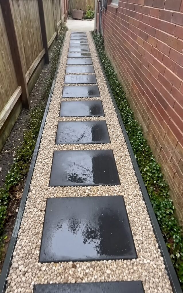 Narrow side-yard walkway with dark rectangular stepping-stone pavers evenly spaced in light tan gravel, bordered by black landscape edging and flanked by a wooden fence on the left and brick house wall on the right, with moss and ivy ground cover along the edges