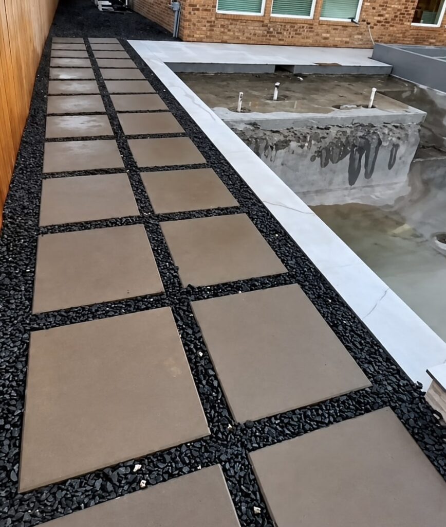 Rooftop or elevated patio walkway with square tan concrete pavers set in dark gravel, beside a concrete retaining wall with white metal trim, overlooking a brick house.