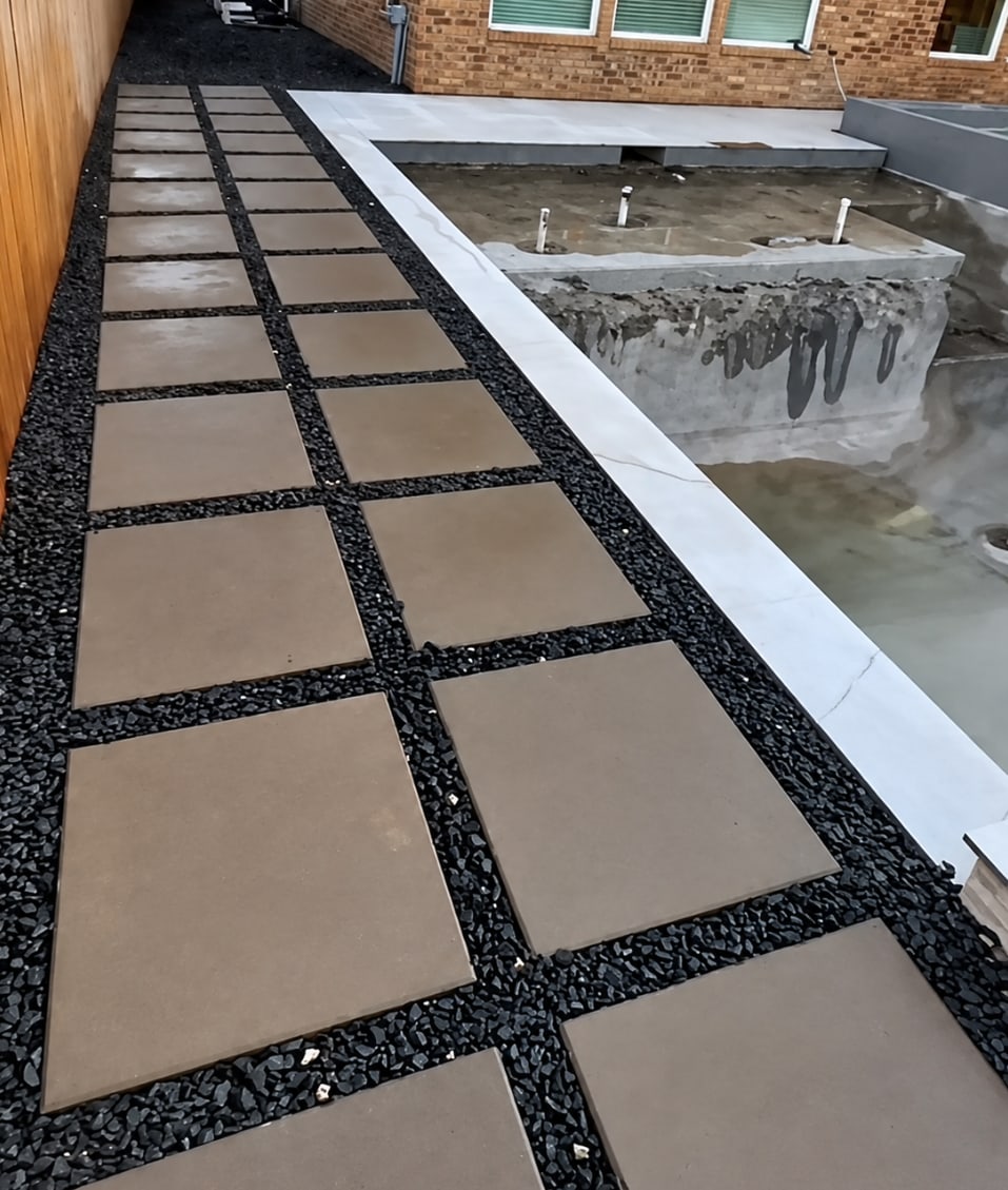 Rooftop or elevated patio walkway with square tan concrete pavers set in dark gravel, beside a concrete retaining wall with white metal trim, overlooking a brick house.