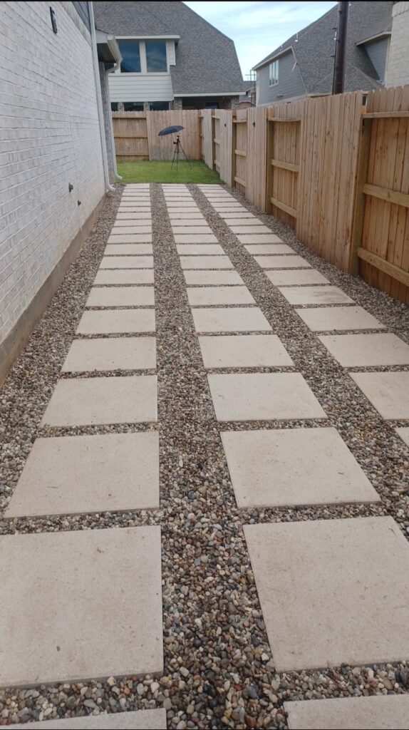 Wide backyard walkway of large rectangular concrete pavers set apart in light gravel, between wooden fences beside a brick house