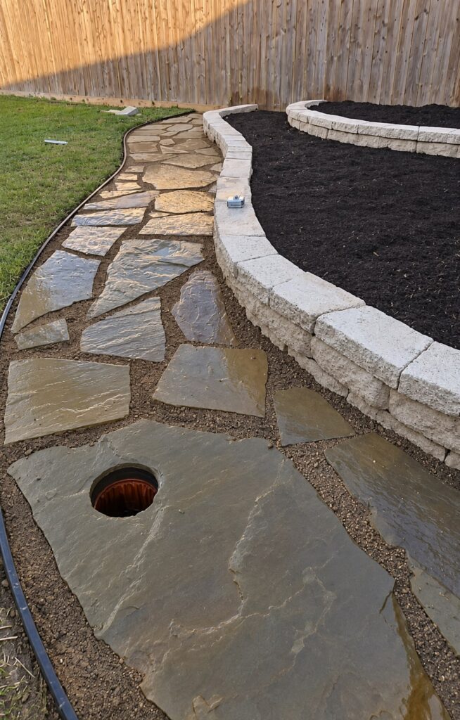 Natural flagstone stepping stones in tan gravel running beside a low block wall, with a wooden fence and planted beds behind