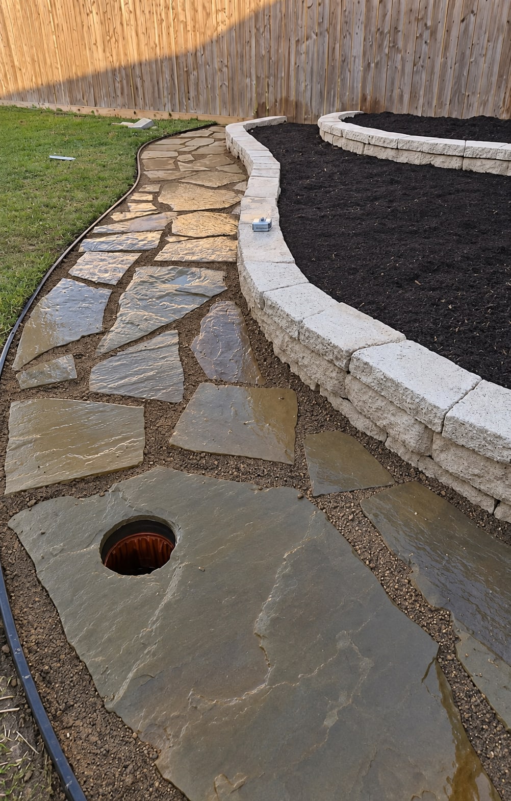 Natural flagstone stepping stones in tan gravel running beside a low tan block wall, with a wooden fence and planted beds behind