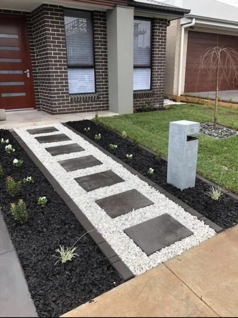 Modern front entry walkway of dark square stepping stones set in light gravel between dark mulch beds, leading to a brick home