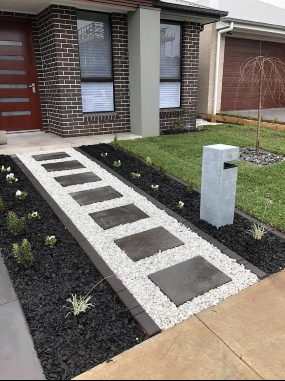Modern front entry walkway of dark square stepping stones set in light gravel, between dark mulch beds, leading to a brick home