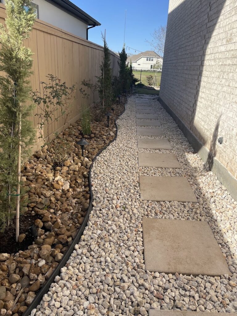 Narrow side-yard walkway of large rectangular concrete stepping stones in light gravel, between a fence with planting beds and a brick house wall